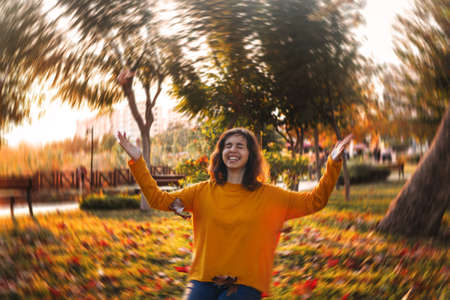 Curly young girl in yellow sweater and jeans sitting on grass and throws up dry leaves.の写真素材