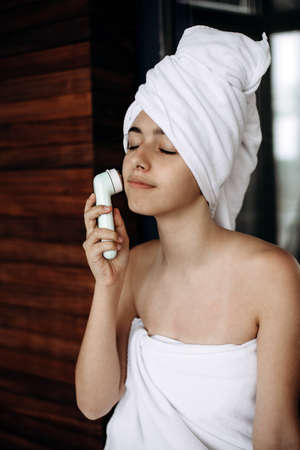 Young girl with towel on head makes facial massage with wireless electric massagerの写真素材