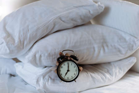 Hand keeps alarm clock with seven o clock on it. Hand surround with pillows on bedの写真素材