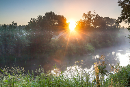Summer landscape with river and sunrise. Foggy scenery with sun sunbeams shine through treesの写真素材