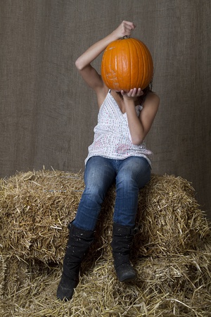 young brunette girl sitting on bales of hay with a pumpkin in front of her faceの写真素材