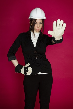 Young woman wearing white hard hat and construction glove, holding her hand up as a stop sign against a pink backgroundの写真素材