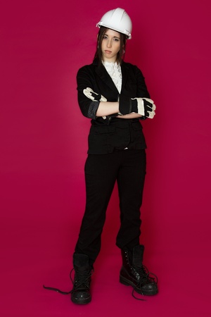 Young woman wearing white hard hat, construction glove and boots that are to big for her with a perplex expression against a pink backgroundの写真素材