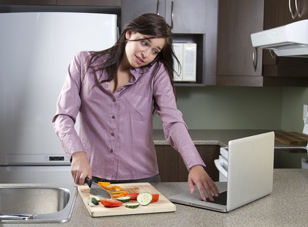 Young woman trying to cut vegetables while working on a laptop and talking on the cellphoneの写真素材