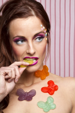 Young woman with colorful makeup and star candy glued to her face and gummy butterfly glued to her body, sucking on the wing of a yellow butterflyの写真素材