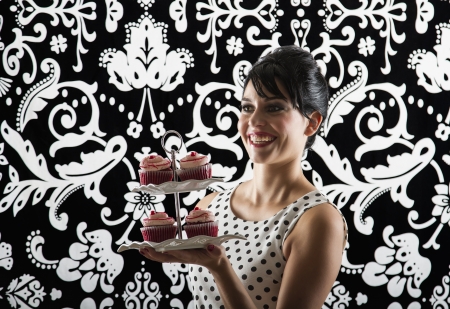 young woman is stylist 60's inspired clothing, with a big smile, holding a plate of velvet cupcakeの写真素材