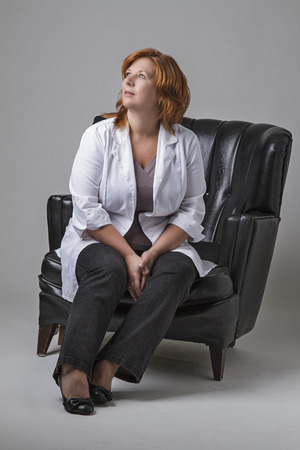 forty year old woman with red hair wearing a doctor lab coat, sitting in a loveseatの写真素材