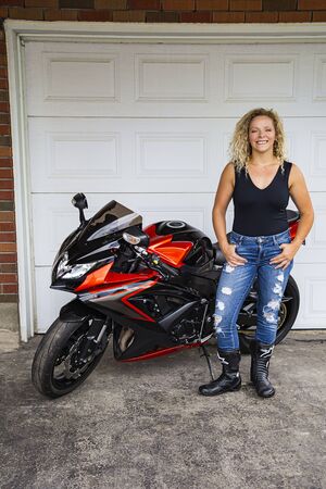 Twenty something blond woman wearing stylish ripped jeans and a black tank top, standing in from of a sport motocycle, with a happy expressionの写真素材