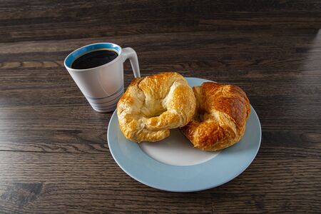 two croissants on a blue rimmed plate with a black coffee against a wood backgroundの写真素材