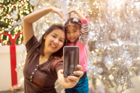 select focus asian mother hand hold mobile with daughter fot take selfie photo with Christmas tree decoration backgroundの写真素材