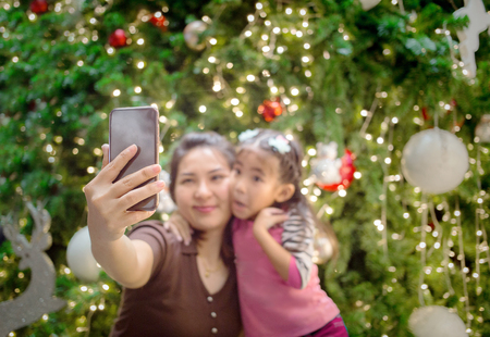 select focus asian mother hand hold mobile with daughter fot take selfie photo with Christmas tree decoration background .Selfue with mobile is behavior of digital generation to capture memmories.の写真素材