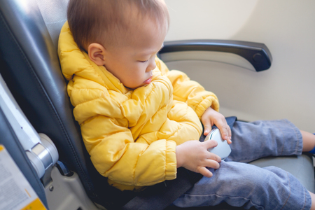 Cute little Asian 20 months / 1 year old toddler baby boy child wearing & fasten seat belts while sitting on airplane seat. Safety measures on board. Precautions on plane during flight for kidの写真素材