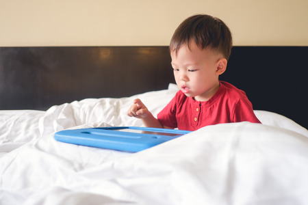 Cute little Asian 2-3 years old toddler boy child sitting in bed watching a video from tablet pc. Kids playing with tablet computer, Gadget-addicted children, pseudo-autism concept - Selective focusの写真素材