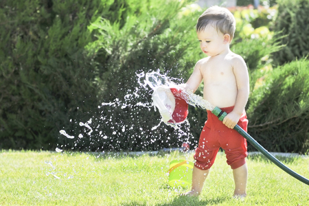 Little boy pours from a hose in the garden on a hot summer day on a green lawn, splashing waterの写真素材