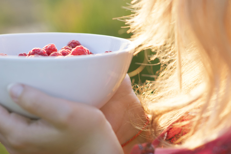 Young girl holding a plate of raspberries, sitting on green grass, summer, dessert