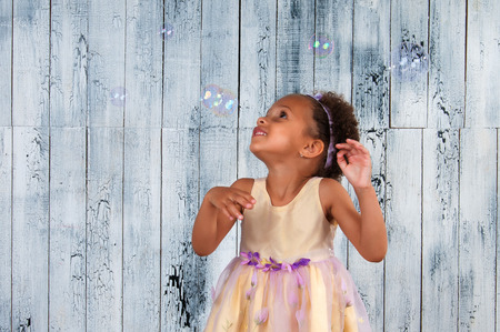 Happy smiling african  girl blowing soap bubbles on the background of wooden wallの写真素材