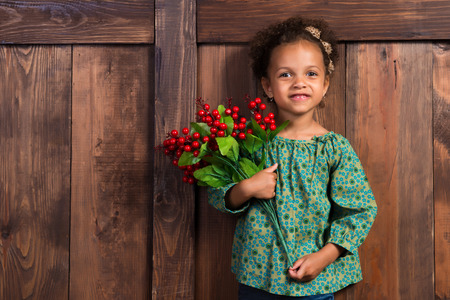 Smiling little african  girl in rural shirt with bunch of berries on  background of brown wooden wallの写真素材