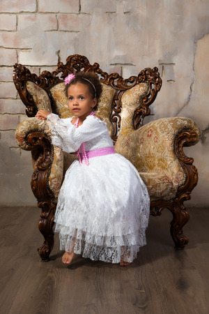 Cute african  girl with curly hair in a white lace dress on vintage chairの写真素材