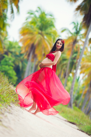 Beautiful pregnant girl in red  dress on sandy beach with palm treesの写真素材