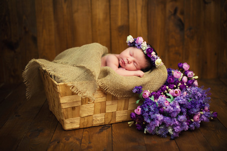 Newborn baby girl with a wreath in a wicker basket with a bouquet of purple wild flowersの写真素材