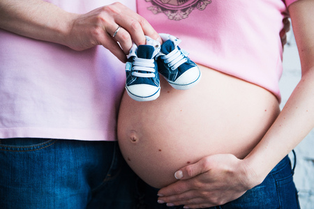 Childrens shoes in the hands of a young dad on the background of the belly of a pregnant womanの写真素材