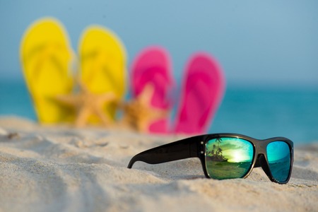 Glasses with yellow and pink sandals stand in the sand against the background of the seaの写真素材