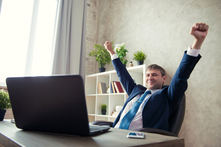 Happy excited young businessman sitting on workplace and celebrating success.の写真素材