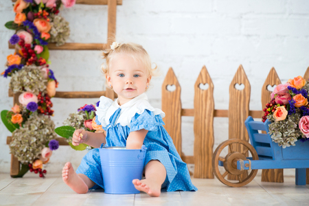 Small toddler lovely girl in blue dress with a bucket and a cart of flowers in studio scenery beautiful garden. Little gardener.の写真素材