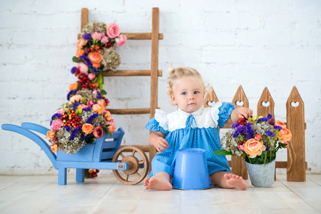 Small toddler lovely girl in blue dress with a bucket and a cart of flowers in studio scenery beautiful garden. Little gardener.の写真素材