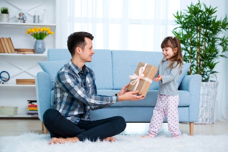 Cute little girl, daughter, sister gives a gift box to young dad father or brother. Both are smiling. Father's day holiday concept, Children's Day.の写真素材