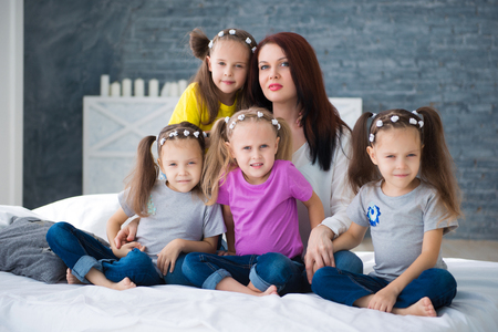 Large friendly family, many children: mom and four pretty cheerful girls triple twins sisters sitting on a bed against a gray brick wall.の写真素材