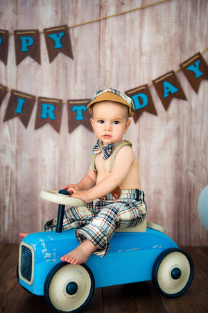 Little boy kid gentleman in retro costume with suspenders and cap is sitting on a wooden car. Children's party with balloons Happy Birthday, 1 year.の写真素材