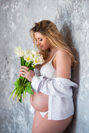 Beautiful young pregnant blonde woman love flowers white tulips. Spring mood, freshness, women's Day, health and beautiful pregnancy.の写真素材