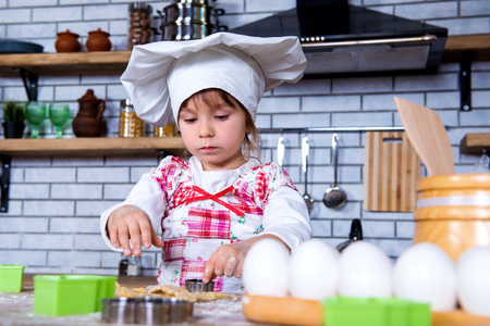 A girl in a chef's hat is cooking gingerbread in the kitchen, making cakes from cookies tins.の写真素材