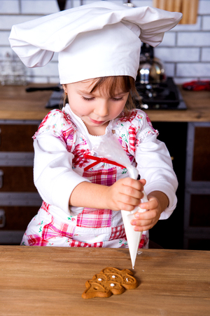 Cute girl in a cooking chef's hat decorate ready-made baked gingerbread Christmas tree figurine with sweet icing and cream.の写真素材