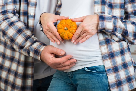 husband and wife in jeans and a plaid shirt holds little pumpkin in their hands. Couple posing a heart with their fingers on belly of expectant mother.の写真素材