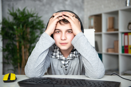 Oops, failure, loss. Teenager boy lost in a computer game. A schoolboy sits upset in front of a computer monitor, covering his face with his hands.の写真素材