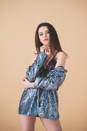 Beautiful sexy brunette girl in a black lace jumpsuit and silver jacket standing on a beige paper background in the studio.の写真素材