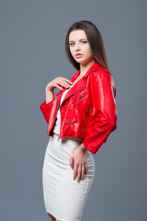 Fashionable style, fashion women's clothing, color combination. Beautiful brunette girl in white dress and red leather jacket isolated gray background.の写真素材