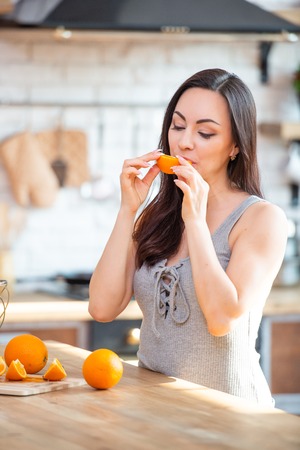 A young woman in a gray t-shirt tries to eat a slice of juicy orange in the interior of the kitchen.の写真素材