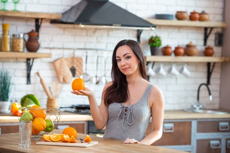 A young woman in a gray t-shirt holds an orange in her hand. Healthy eating concept. In the interior of the kitchen.の写真素材