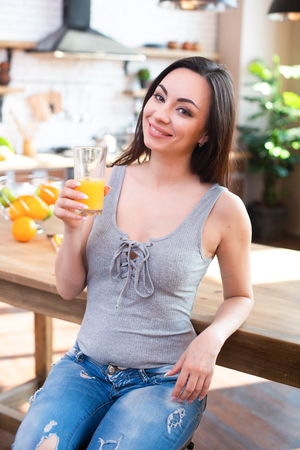 Smiling young woman in gray t-shirt drinks fresh orange juice in kitchen interior.の写真素材