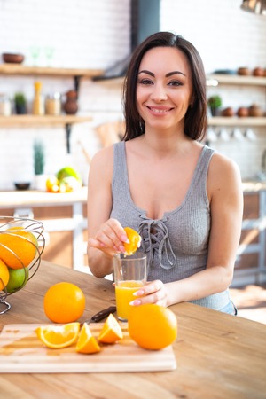 Smiling young woman in a gray T-shirt squeezes a fresh orange on fruit juice. Healthy eating concept. In the interior of the kitchen.の写真素材