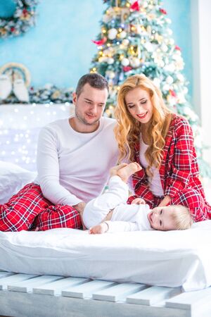 Happy family in plaid pajamas fooling around near the christmas tree. Mother, father and little son laughing on the bed.の写真素材