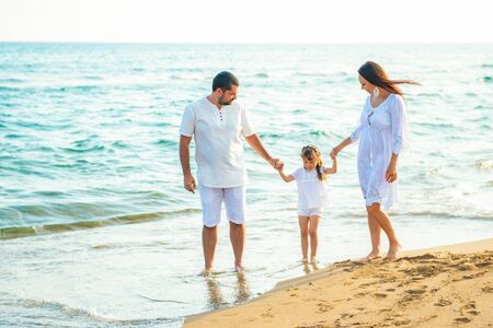 Happy family mom, dad and little daughter walk along the coast on sea beach at resort.