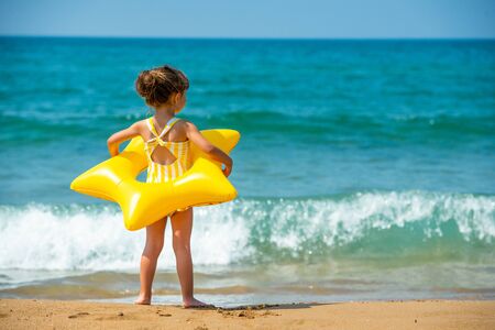 A little child girl 3 years old stands with a yellow swimming ring in the form of a starfish and looks at the sea. Back view.の写真素材