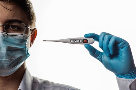 A doctor in a glasses and surgical mask holds an electronic thermometer to measure temperature....