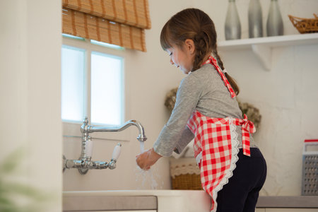 Little girl with pigtails in a culinary apron washes her hands in the kitchen.の写真素材