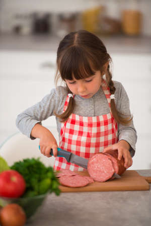 Cute little girl with pigtails in a plaid culinary apron cuts sausage into sandwiches in the kitchen at the table.の写真素材