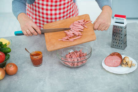 Woman in checkered apron cuts sausage in the kitchen for pizza toppings.